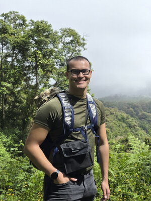 A smiling man in a t-shirt, wearing a backpack, standing in front of trees and green countryside