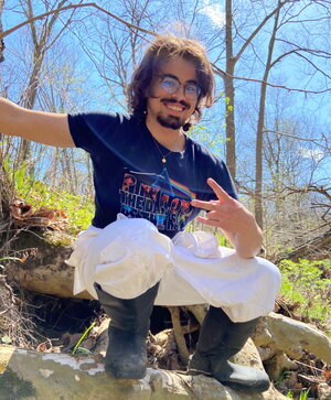 A man squatting outside in a blue shirt giving the peace sign outside