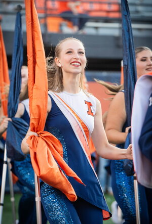 A smiling woman in the Illini color guard outfit holding a flag