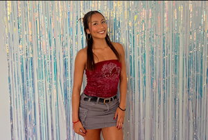 A smiling woman in a red shirt and grey skirt standing in front of a wall with blue tinsel