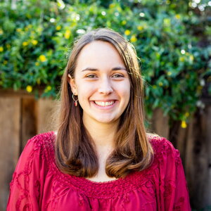 A smiling woman with long hair in a reddish-pink shirt standing outside in front of a fence