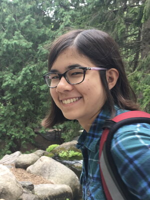 A smiling woman in glasses wearing a backpack and blue plaid shirt outside