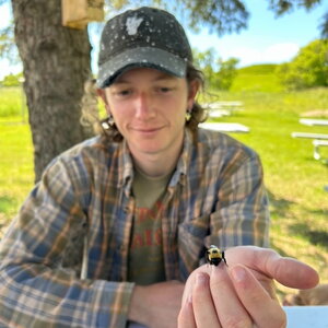A man in a hat and plaid shirt holding a bee to the camera