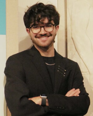A smiling brunette man in glasses wearing all black with his arms crossed