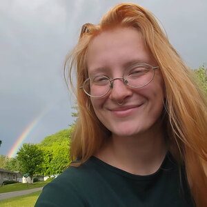 A smiling redheaded woman in round glasses standing in front of a rainbow