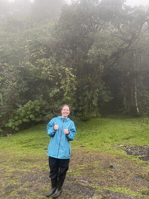 A smiling woman in a blue rain coat giving thumbs up outside in the rain