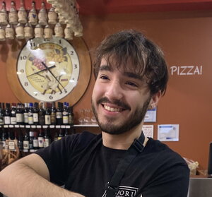 A smiling bearded man in an italian business with a clock and wine in the background