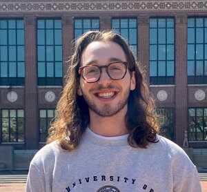 A smiling man with long hair in round glasses and a sweater standing in front of a brick building
