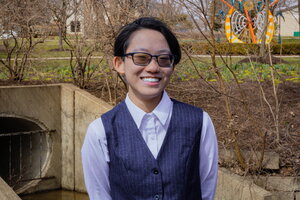 A smiling man wearing glasses in dark vest and purple button up standing outside