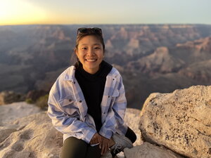 A smiling woman in a blue and white jacket sitting on a mesa with mesas behind her