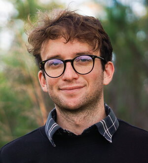A smiling man in round black glasses wearing a sweater with a collar outside