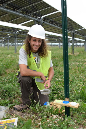 A man with long hair in a hard hat and neon vest kneeling outside