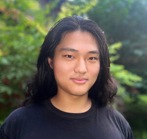 A semi-smiling person with long black hair in a black shirt standing in front of green plants