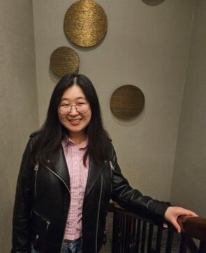 A woman in a leather jacket and pink shirt smiling on a stairwell