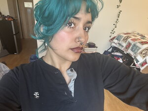 A woman with turquoise hair and black shirt taking a selfie in a bedroom