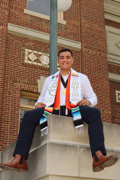 a photo of Ben sitting on a ledge in nice clothes wearing his graduation sash