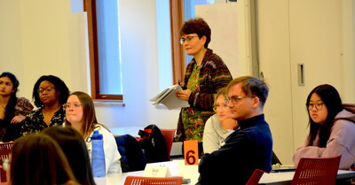 A photo of a person holding papers with students around them.