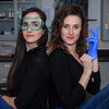 Two women sitting on a lab table. Both are wearing black with jeans. The one on the left is wearing safety goggles, the one on the right is putting on a blue glove.