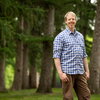 A man in a blue plaid shirt and brown pants standing in front of trees