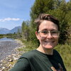 a woman standing next to a river with mountains in the background