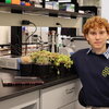 a redheaded man in a blue sweater with a collared shirt underneath sitting in a lab with plants next to him