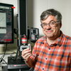 A man smiling in an orange and blue button up in a lab holding an object