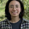A smiling woman in round glasses and a plaid shirt standing outside