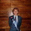 A smiling man in a suit wearing a sash standing in front of a wooden wall