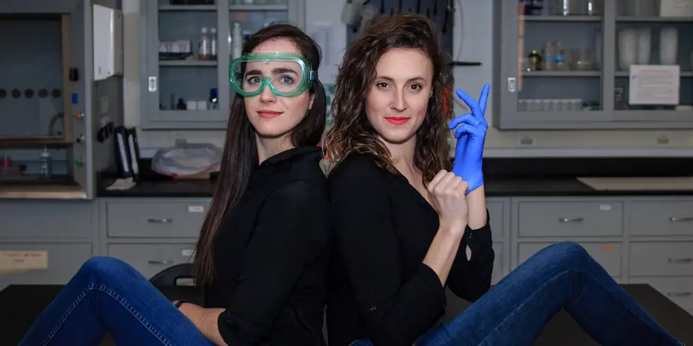 Two women sitting on a lab table. Both are wearing black with jeans. The one on the left is wearing safety goggles, the one on the right is putting on a blue glove.