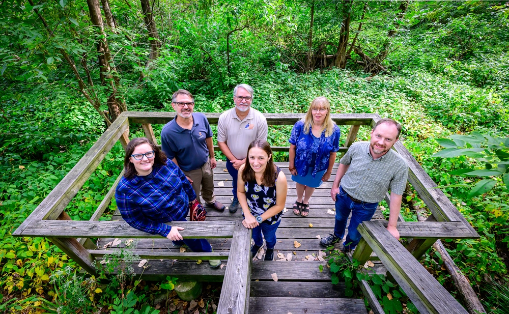 A group of people standing on a wooden platform with woods behind them