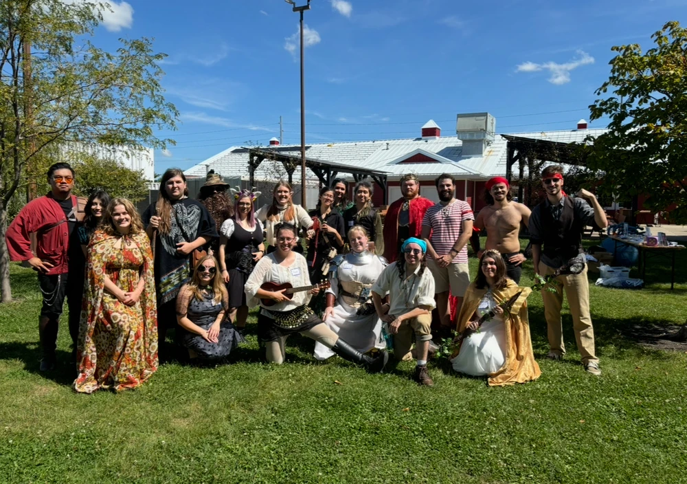 A group of people standing and kneeling in grass wearing fantasy cosplay, with a building behind them