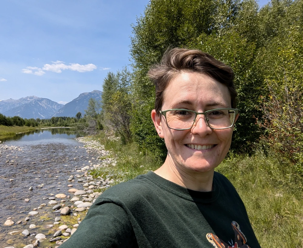 a woman standing next to a river with mountains in the background