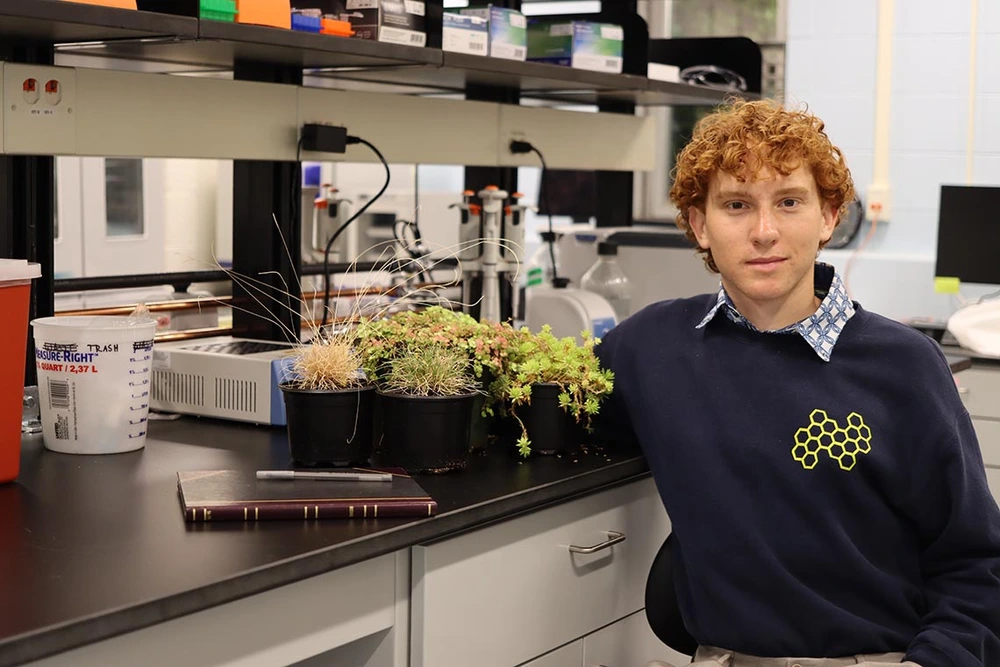 a redheaded man in a blue sweater with a collared shirt underneath sitting in a lab with plants next to him