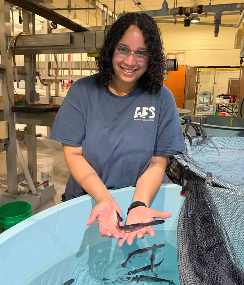 A smiling woman holding a small paddlefish over a large tub with others swimming inside