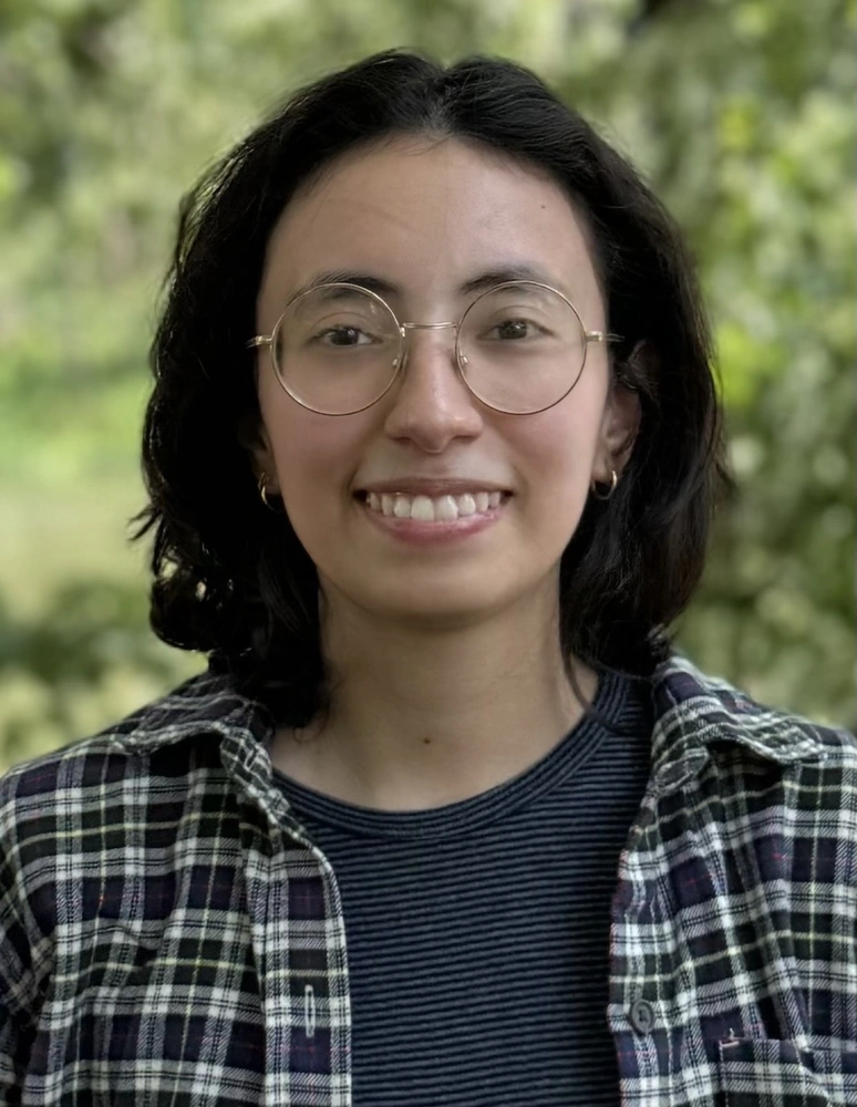 A smiling woman in round glasses and a plaid shirt standing outside