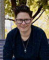 A person wearing glasses in a dark shirt standing in front of a tree