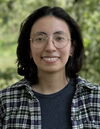 A smiling woman in round glasses and a plaid shirt standing outside