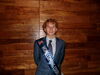 A smiling man in a suit wearing a sash standing in front of a wooden wall
