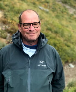 A man wearing glasses in a green jacket standing next to a grassy hill