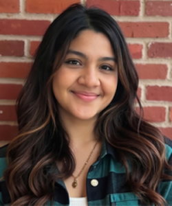 A smiling woman in a green and black flannel standing in front of a brick wall