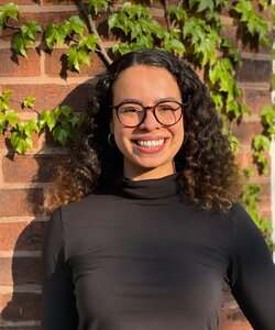 A smiling woman standing in front of a brick wall with plants growing on it