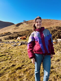 A student in a purple and pink jacket standing on a brown hill with a cow in the background